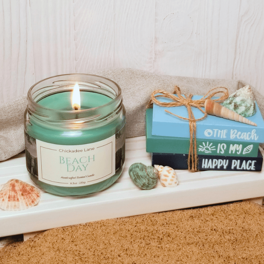 Beach day candle sitting on a white shelf with seashells and sand in front.