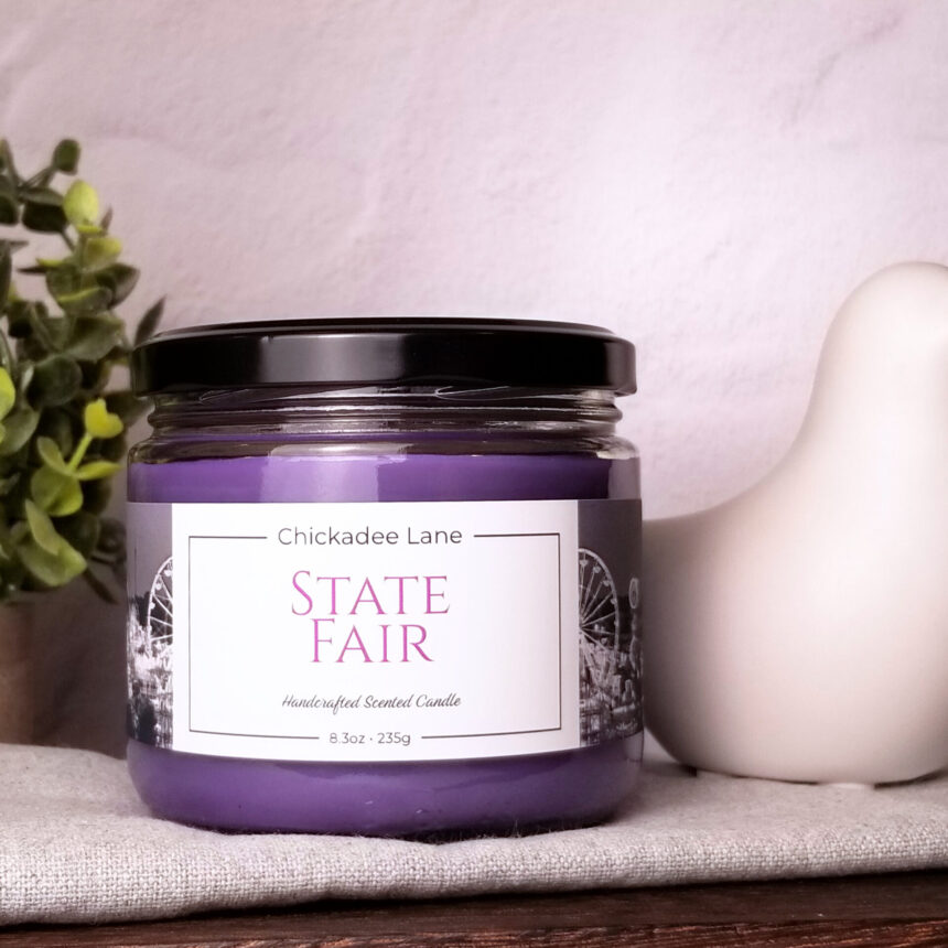 State Fair candle sitting on a shelf between a plant and a porcelain bird.