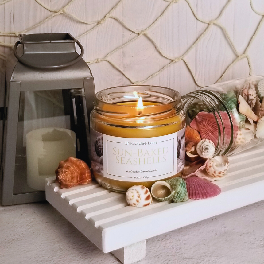Sun Baked Seashells candle sitting on a white shelf with small seashells scattered around it.